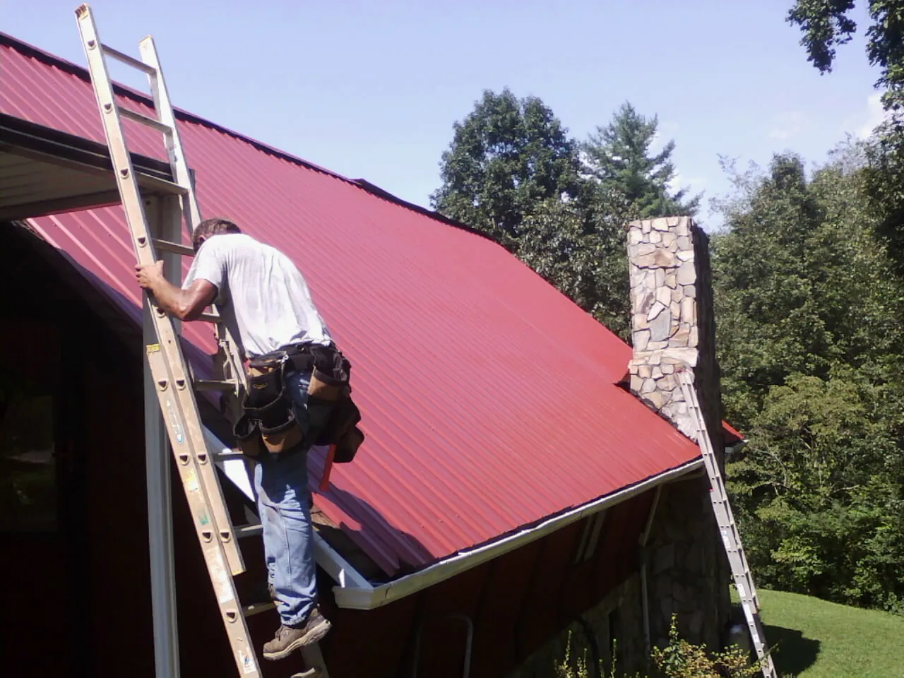 Roofer on a ladder beside a red metal roof and stone chimney — Soffit & Fascia Repair by Roofing Brothers in Gainesville